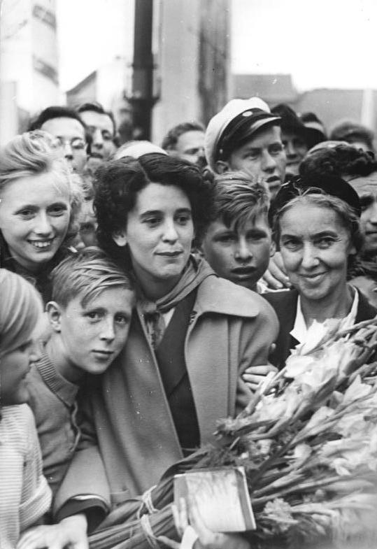 Cette photographie en noir et blanc montre Raymonde Dien au milieu d'une foule. Au centre de l'image, elle porte un manteau et ses cheveux sont sombres. Elle est entourée de trois enfants dont l'un repose sa tête sur son épaule. Elle tient un bouquet de fleurs.