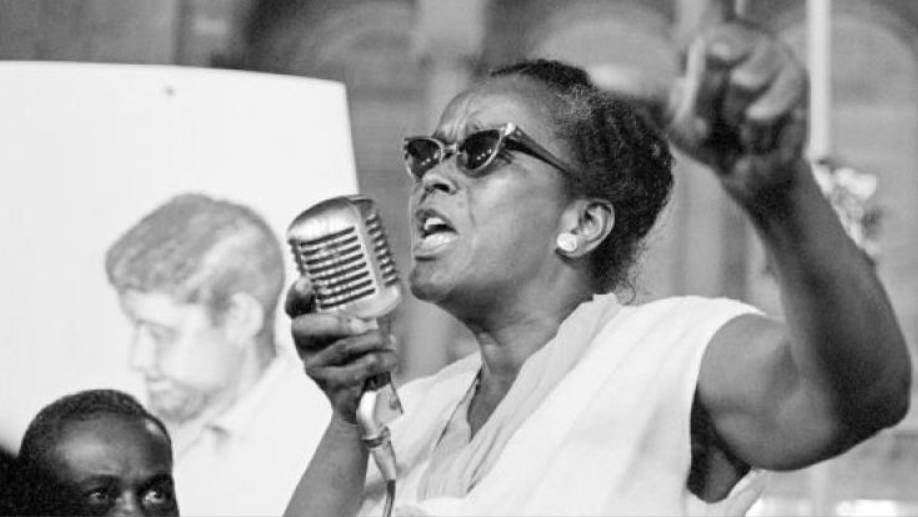 Cette photographie en noir et blanc montre Ella Baker en train de faire un discours lors d'un événement. Elle tient un micro dans sa main gauche et lève sa main droite en l'air. Elle porte des lunettes de soleil. En bas à gauche de l'image, on voit le haut du visage d'un homme afro-américain qui la regarde.