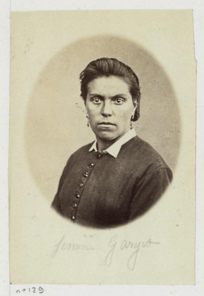 Cette image est une photographie en noir et blanc de Victorine Gorget, prise à la prison des chantiers de Versailles. Elle porte une robe sombre boutonnée sur le devant avec un col blanc. Ses cheveux sont coiffés vers l'arrière. Elle porte des boucles d'oreille et a une expression sévère.