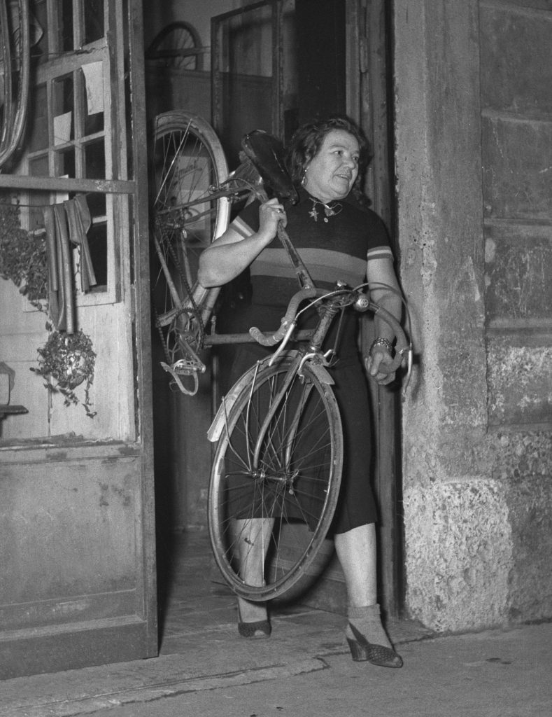 Cette photographie en noir et blanc montre Alfonsina Strada sortant de son magasin de cycles en portant un vélo par le cadre et le guidon, calé sur son épaule gauche.