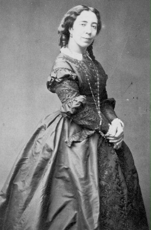 Cette image est une photographie de Pauline Viardot. Debout, elle est vêtue d'une élégante robe sombre et d'un long collier. Bras le long du corps, ses mains sont jointes. Ses cheveux sont longs et coiffés en tresse. Tournée vers la droite, elle regarde vers l'objectif et sourit à demi.